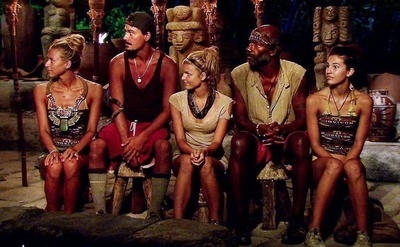 Five contestants on Survivor sitting on a wooden bench with wooden poles and statues decoratively in the background 