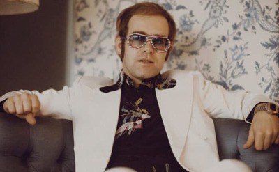 Elton John, wearing a white suit, black shirt with flower motif and multicolored sunglasses.