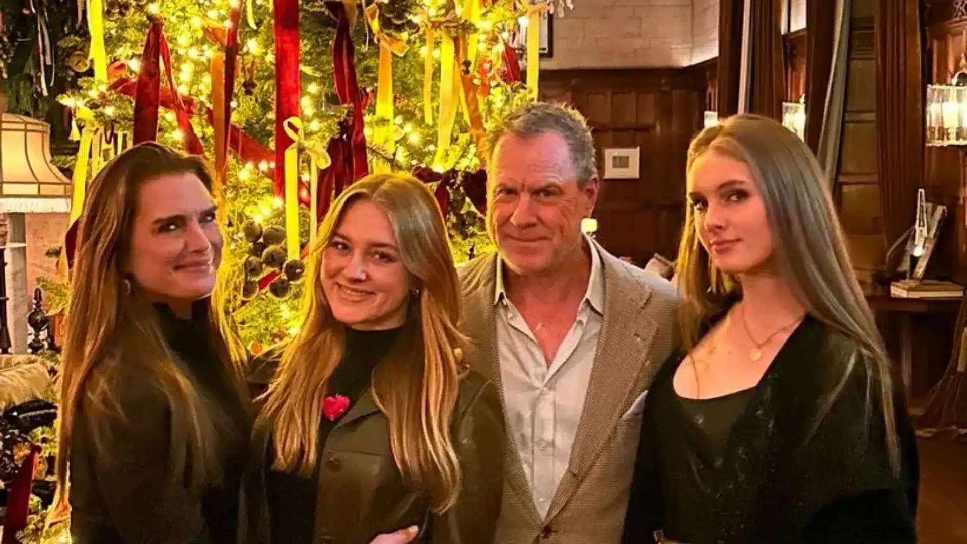 Brooke Shields and her family pose smiling indoors beside a brightly lit Christmas tree decorated with red and yellow ribbons