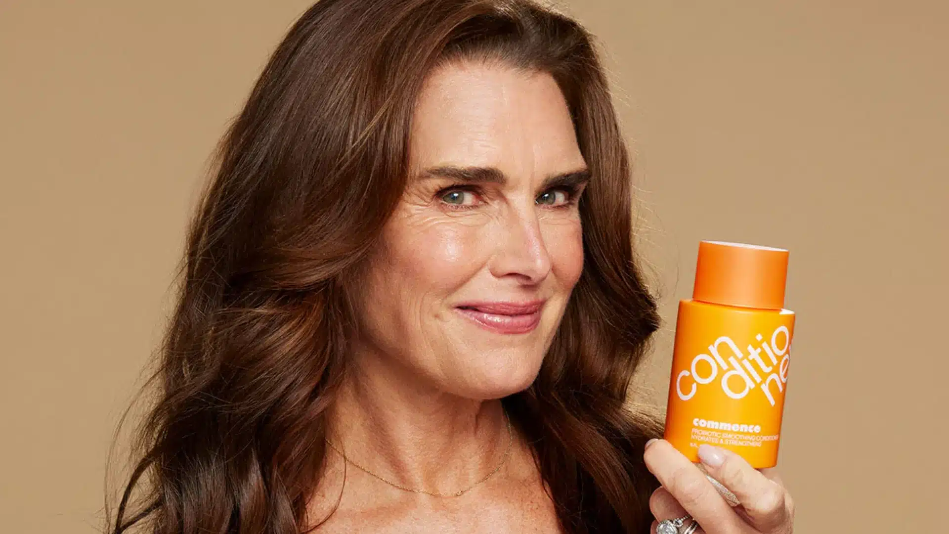 Brooke Shields smiles while holding an orange bottle of conditioner from her brand Commence against a plain tan background