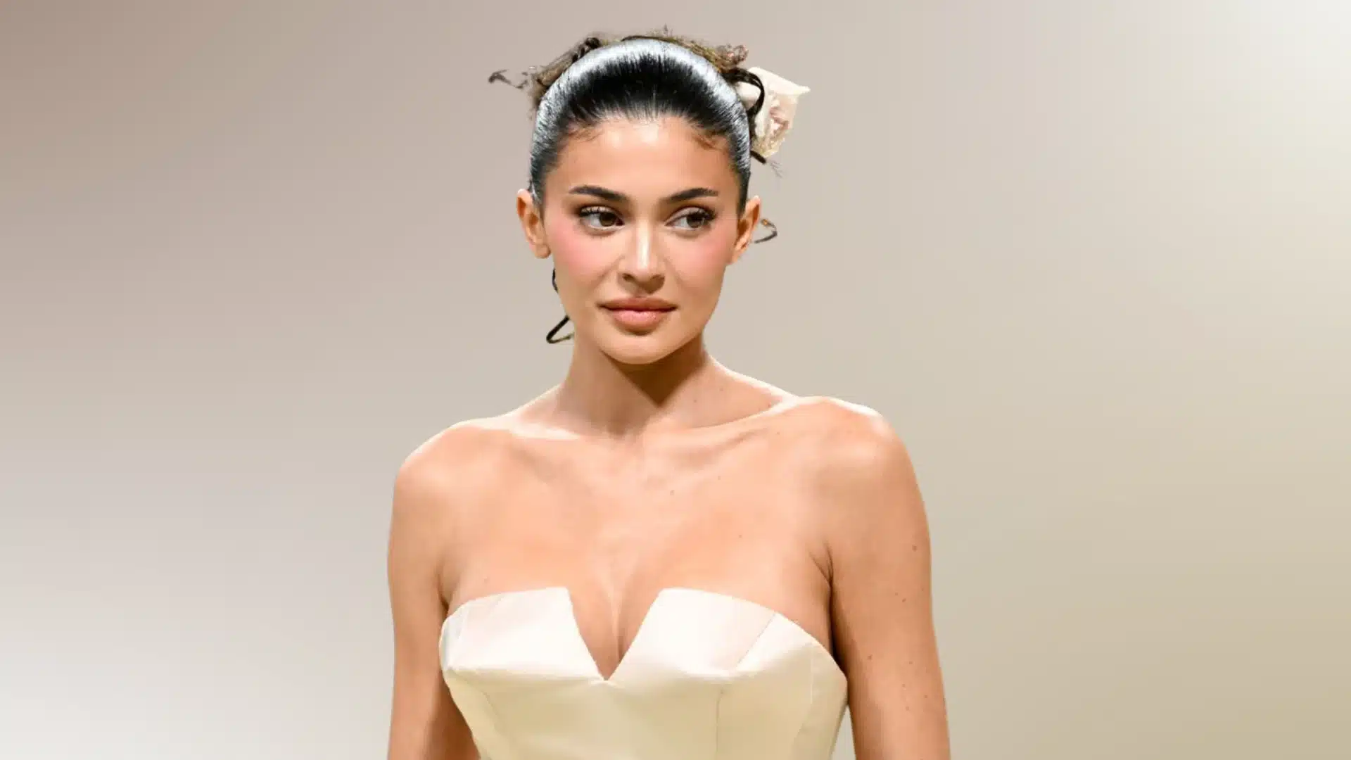 An image of Kylie Jenner wearing a strapless ivory dress with a deep V neckline against a muted beige background