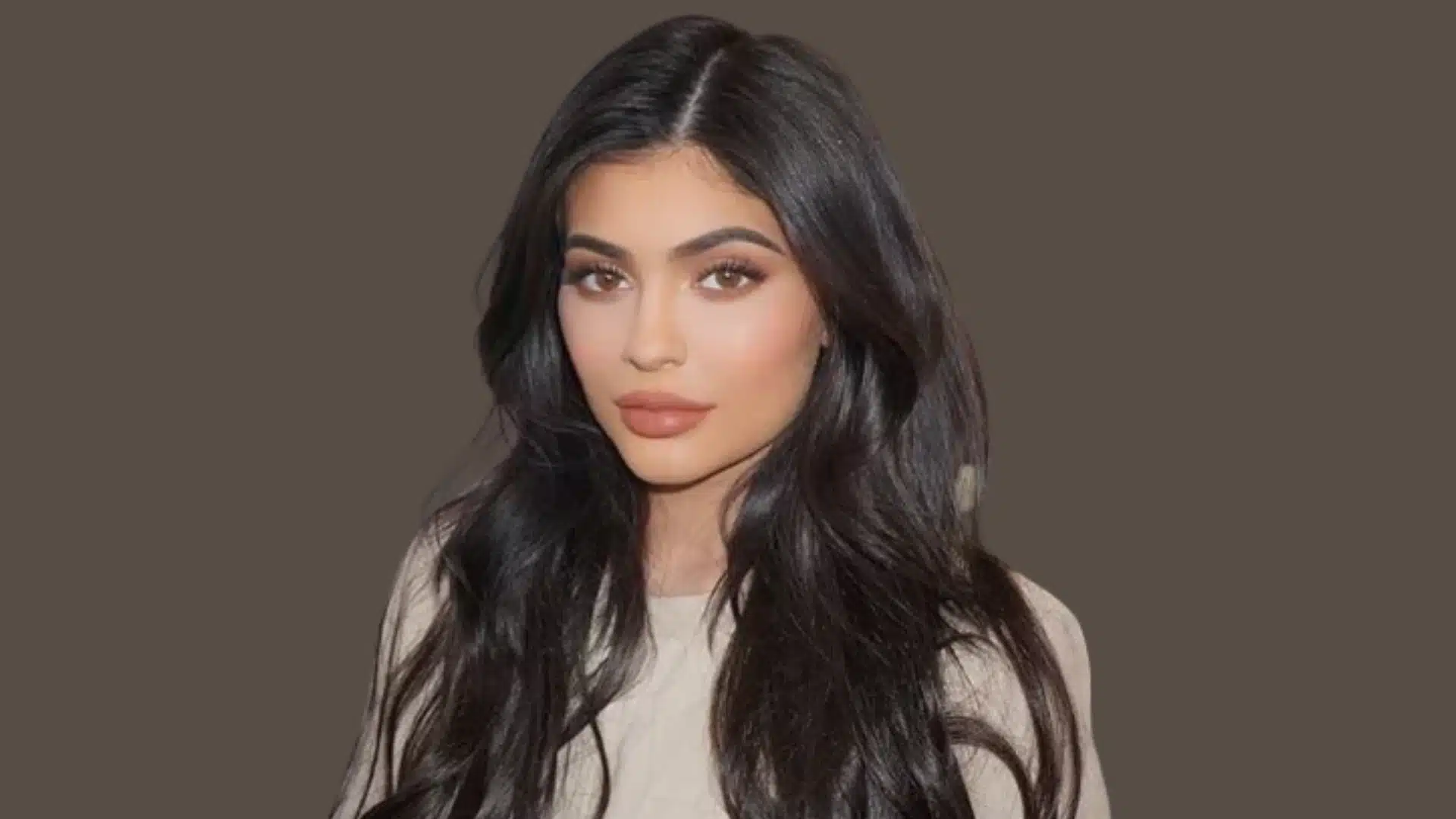 An image of Kylie Jenner with long, dark, wavy hair with a bold, refined look against a solid taupe background