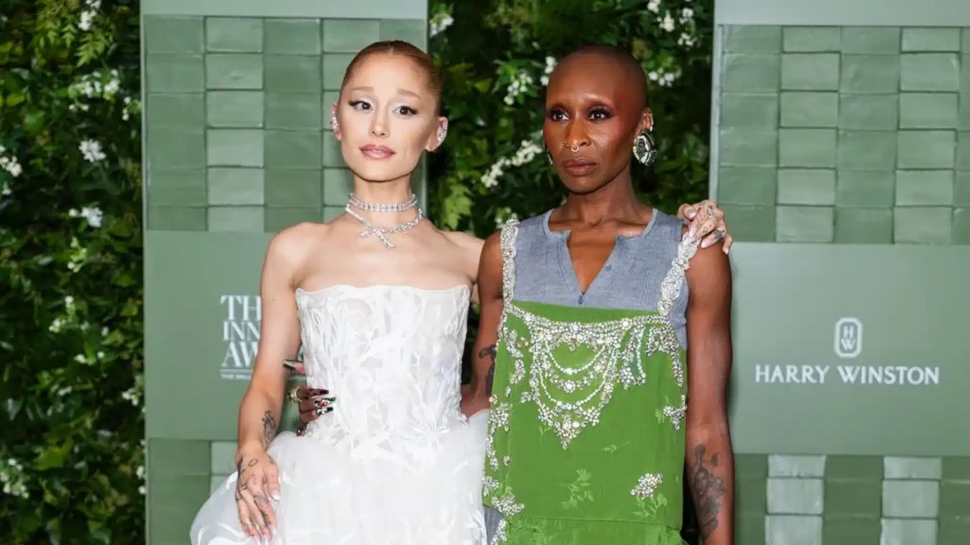 Ariana Grande, in a white gown, and Cynthia Erivo, in a green beaded dress, together at an event with a green backdrop