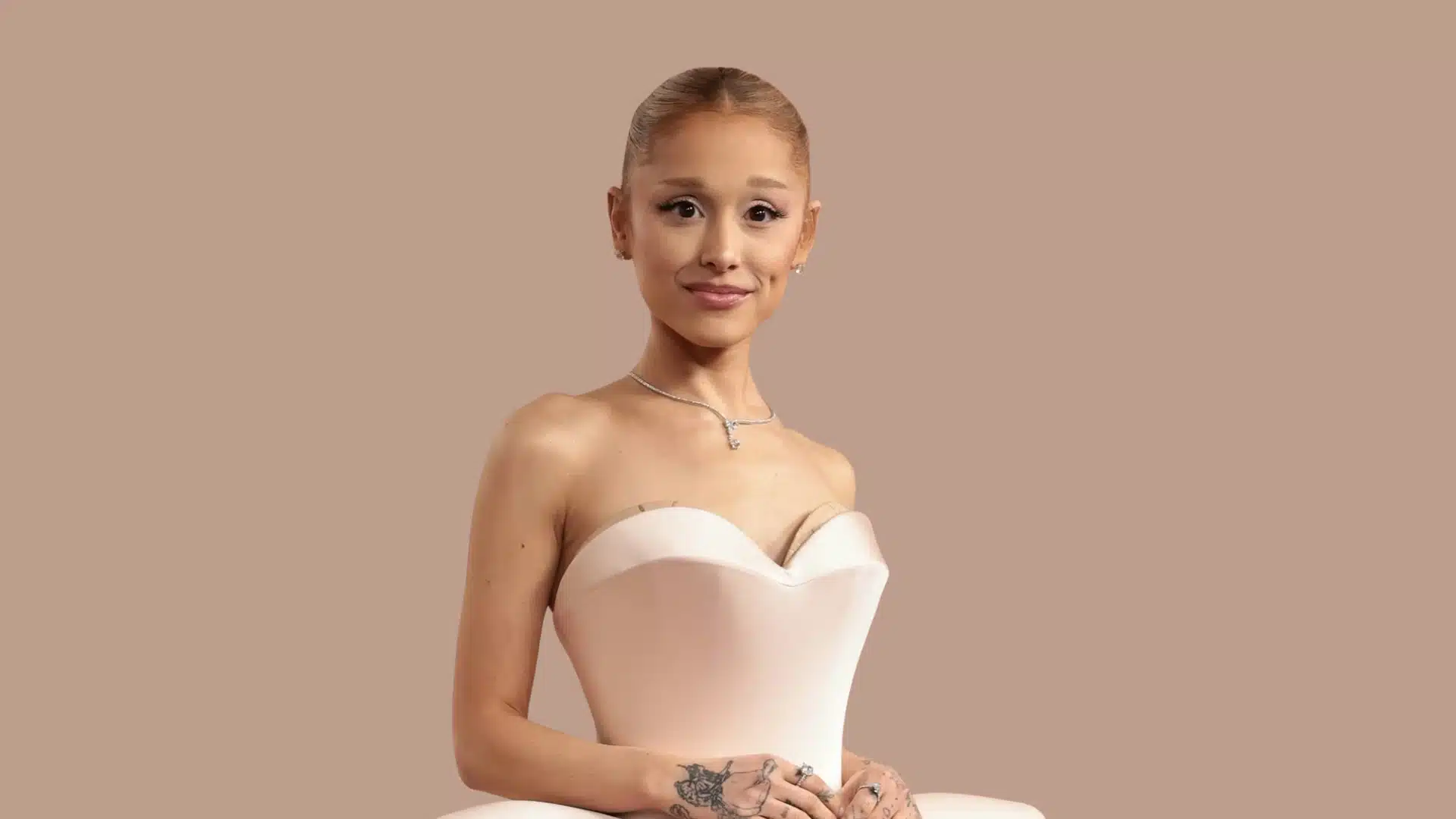 Ariana Grande is in a strapless pale pink gown with her hair pulled back, smiling against a muted beige background