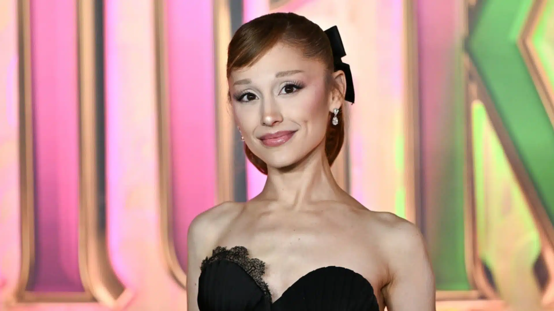 Ariana Grande looks sick while she smiles slightly in a black strapless dress with lace trim and a black bow in her hair