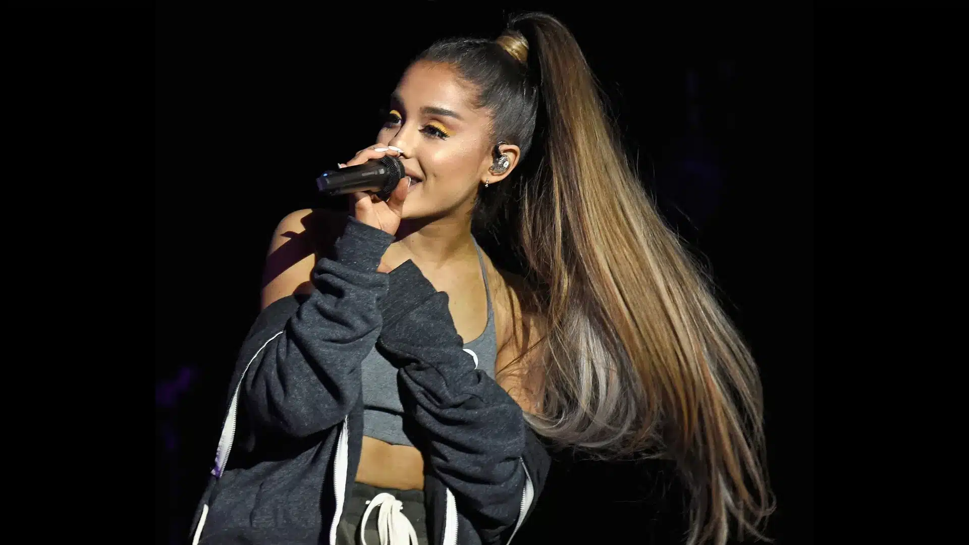 Ariana Grande performing on stage in 2017, holding a microphone, with her signature long, high ponytail, yellow eyeliner and high ponytail