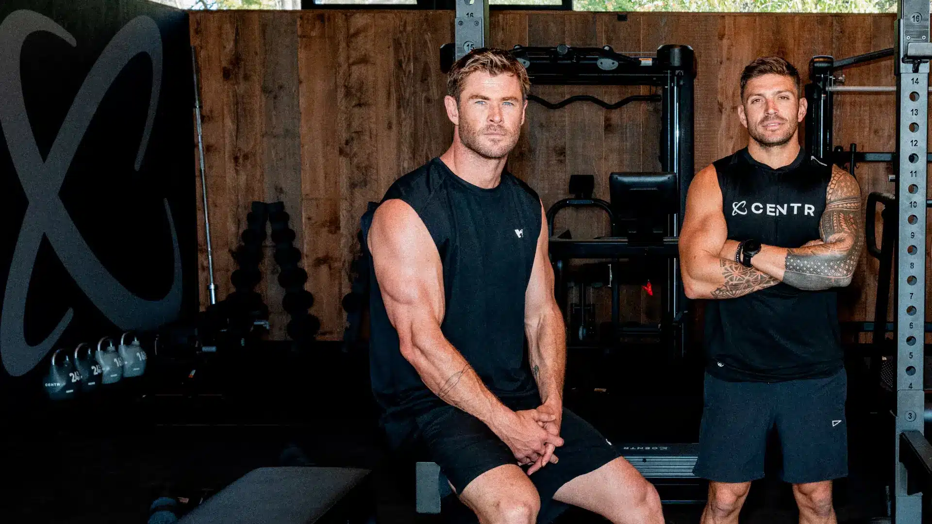 Chris Hemsworth and Luke Zocchi, both in black athletic wear, are at a wood-paneled home gym featuring CENTR branding and workout equipment