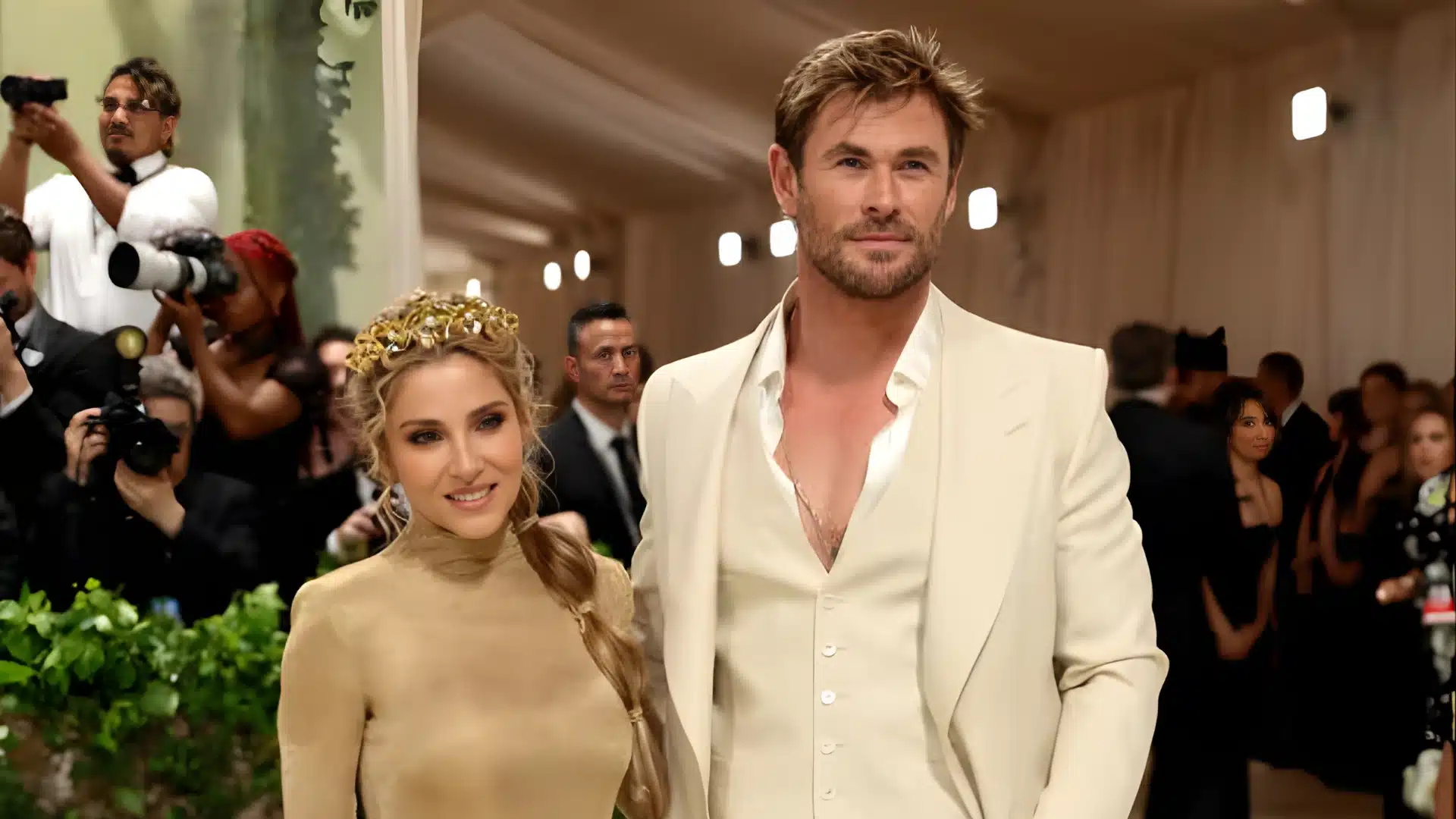 Chris Hemsworth in a cream suit and Elsa Pataky wearing a gold dress pose on a red carpet while surrounded by photographers