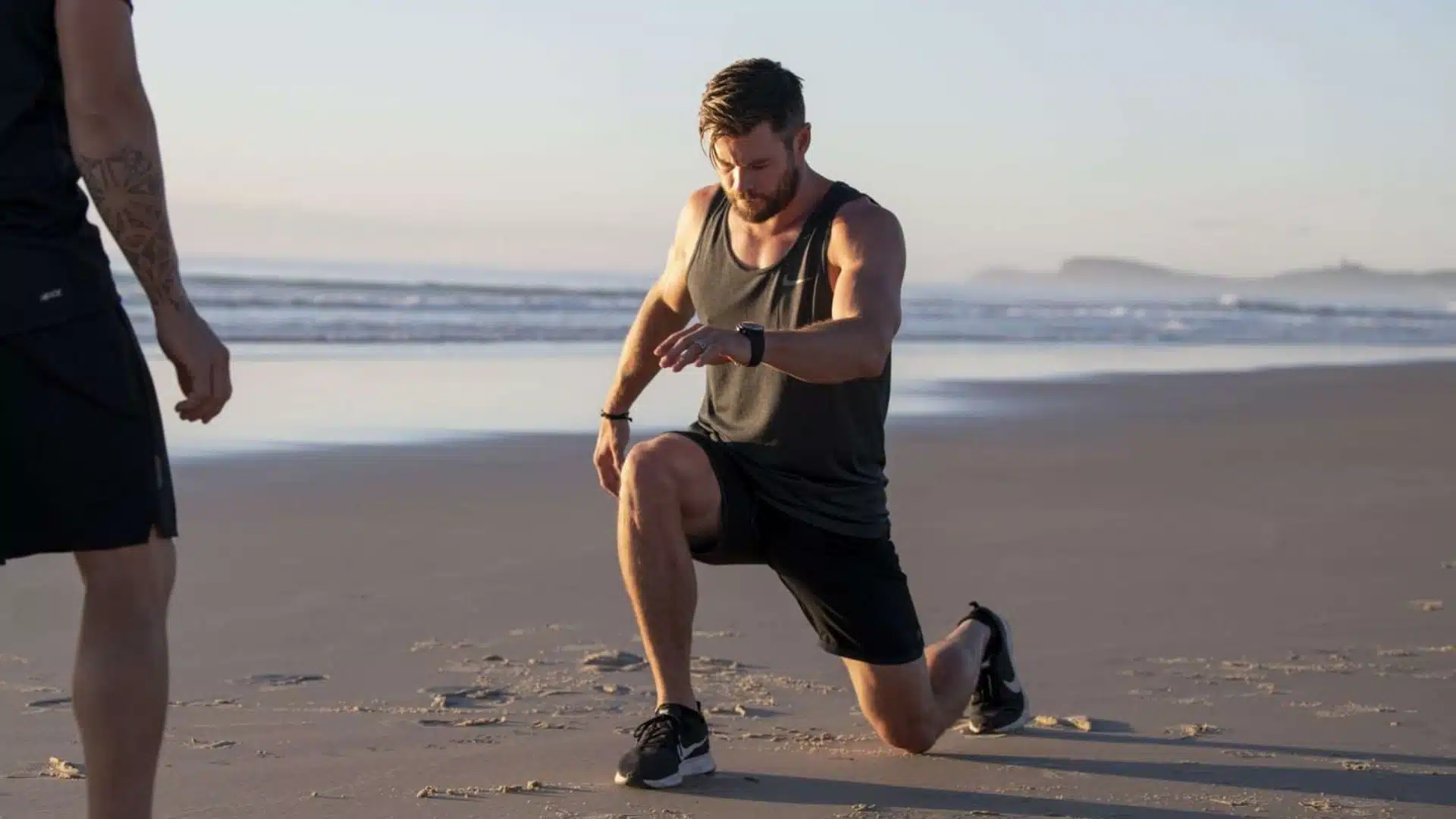 Chris Hemsworth in a tank top, performing a lunge exercise on a sandy beach at sunrise