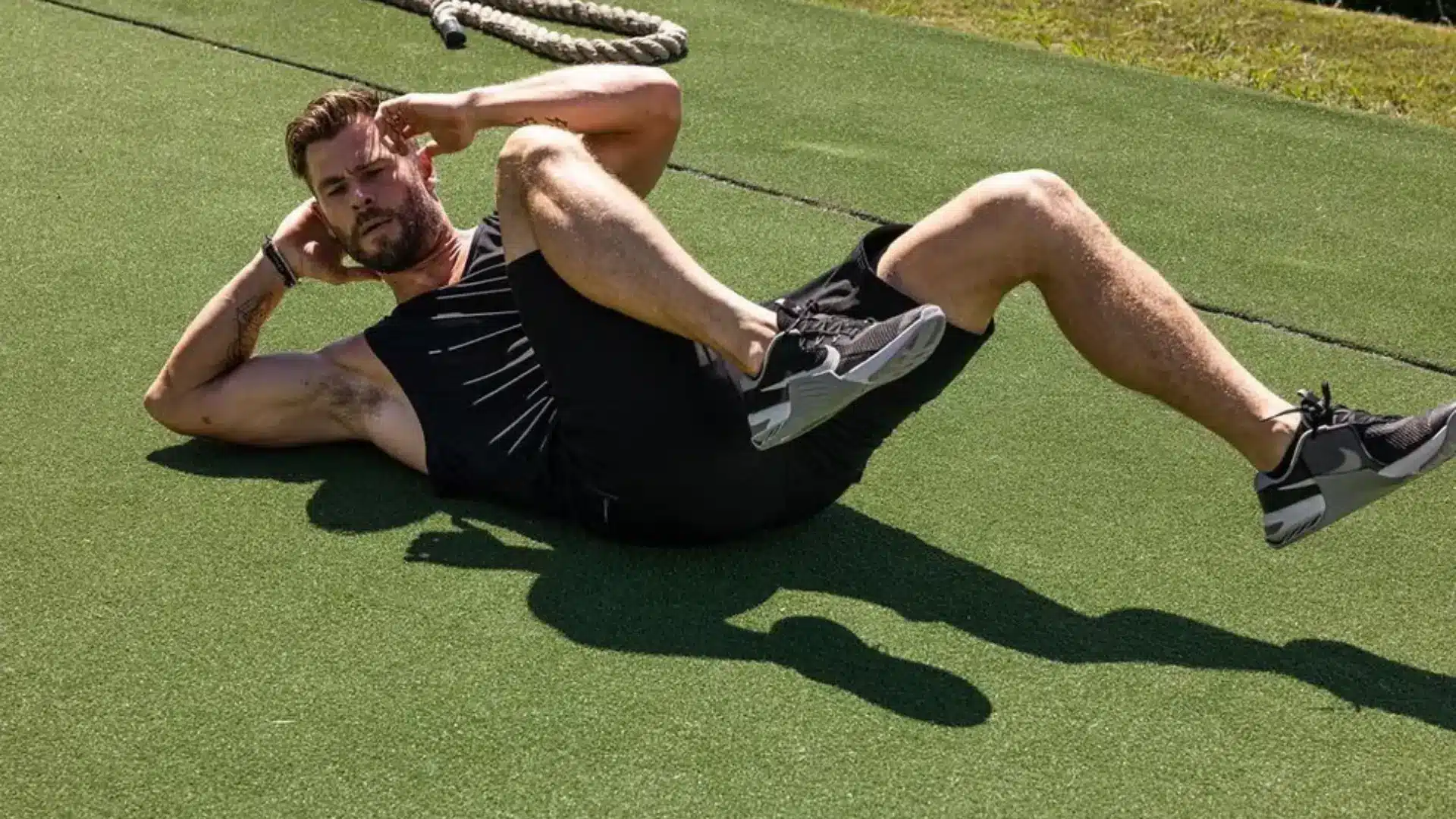 Chris Hemsworth in black workout gear performing a crunch exercise outdoors on green artificial turf