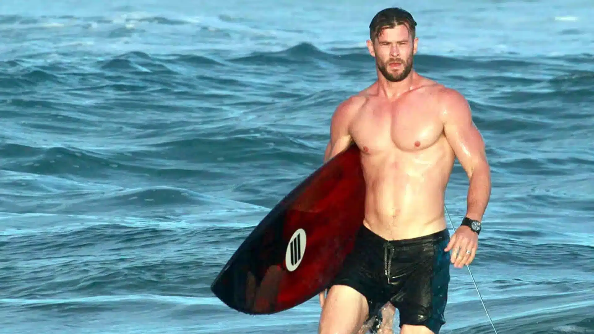 Chris Hemsworth shirtless with a beard, holding a red and black surfboard while wading through the blue ocean waves