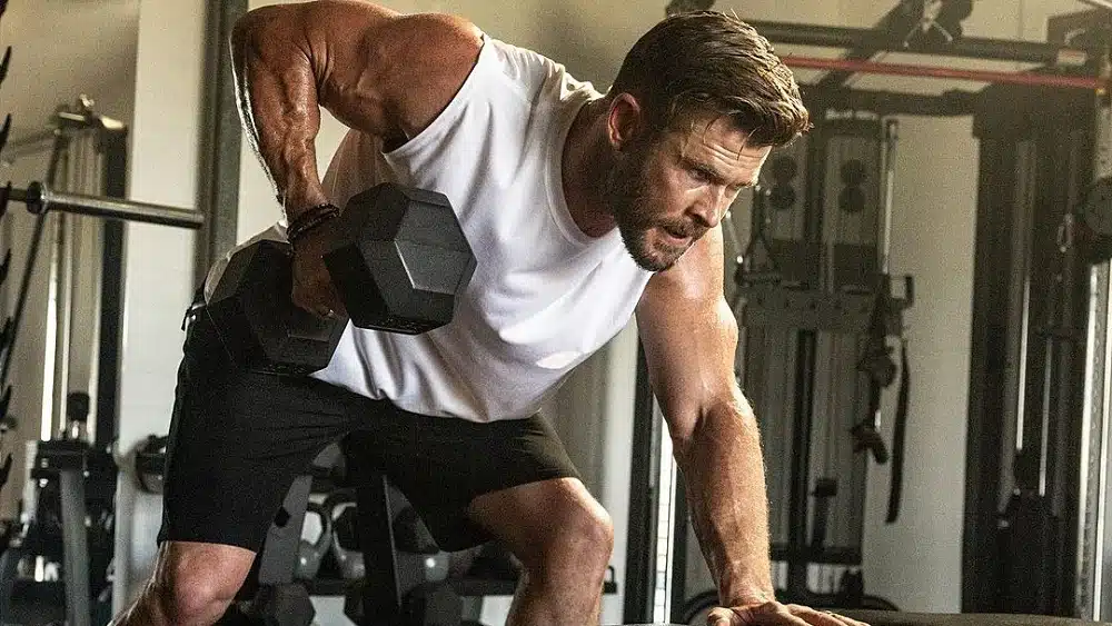 Chris Hemsworth with a beard performing a heavy dumbbell row exercise in a dimly lit gym wearing a white tank top