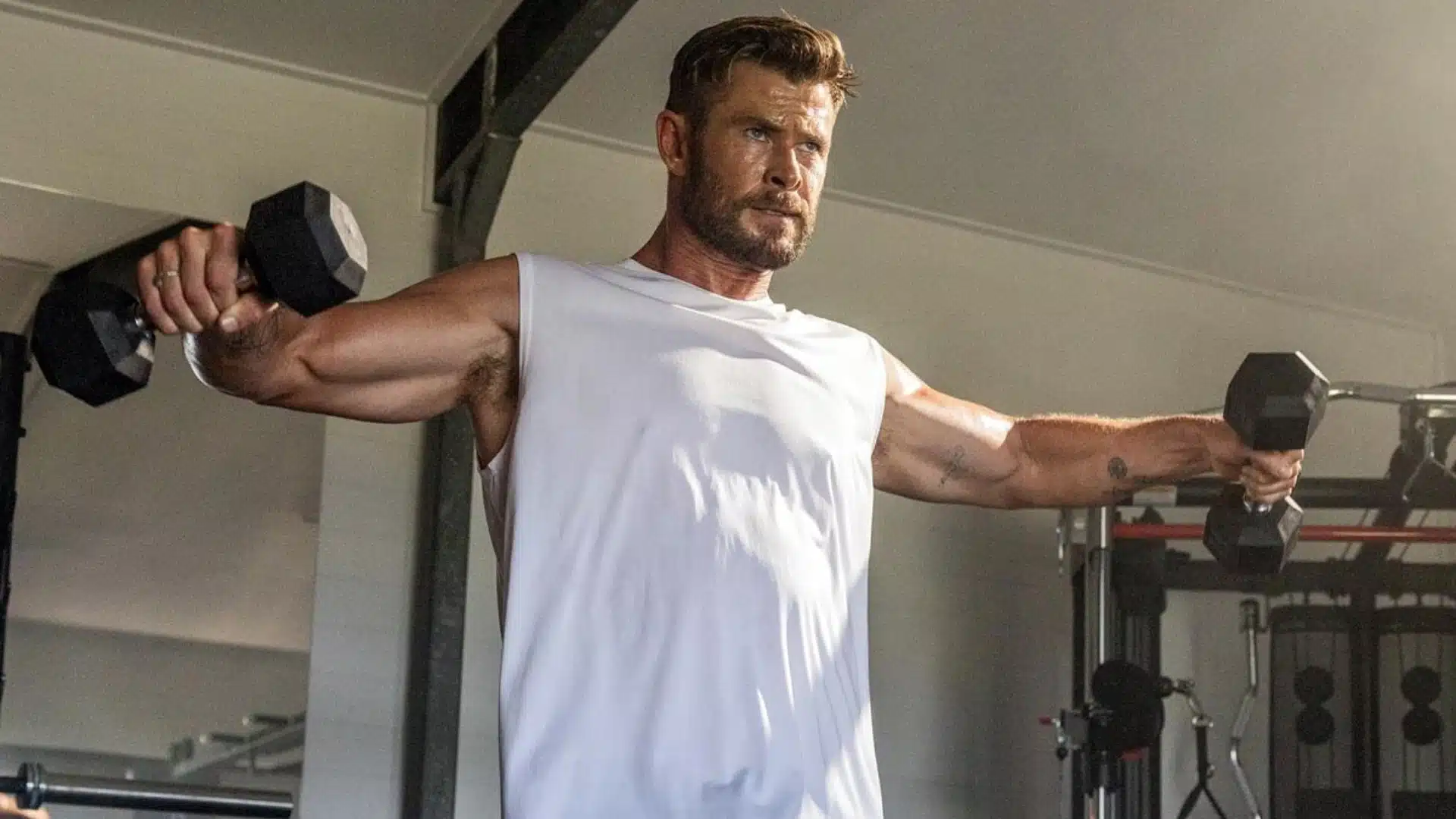 Chris Hemsworth, with a beard, wearing a white tank top, performs a lateral raise exercise holding black dumbbells in a gym setting