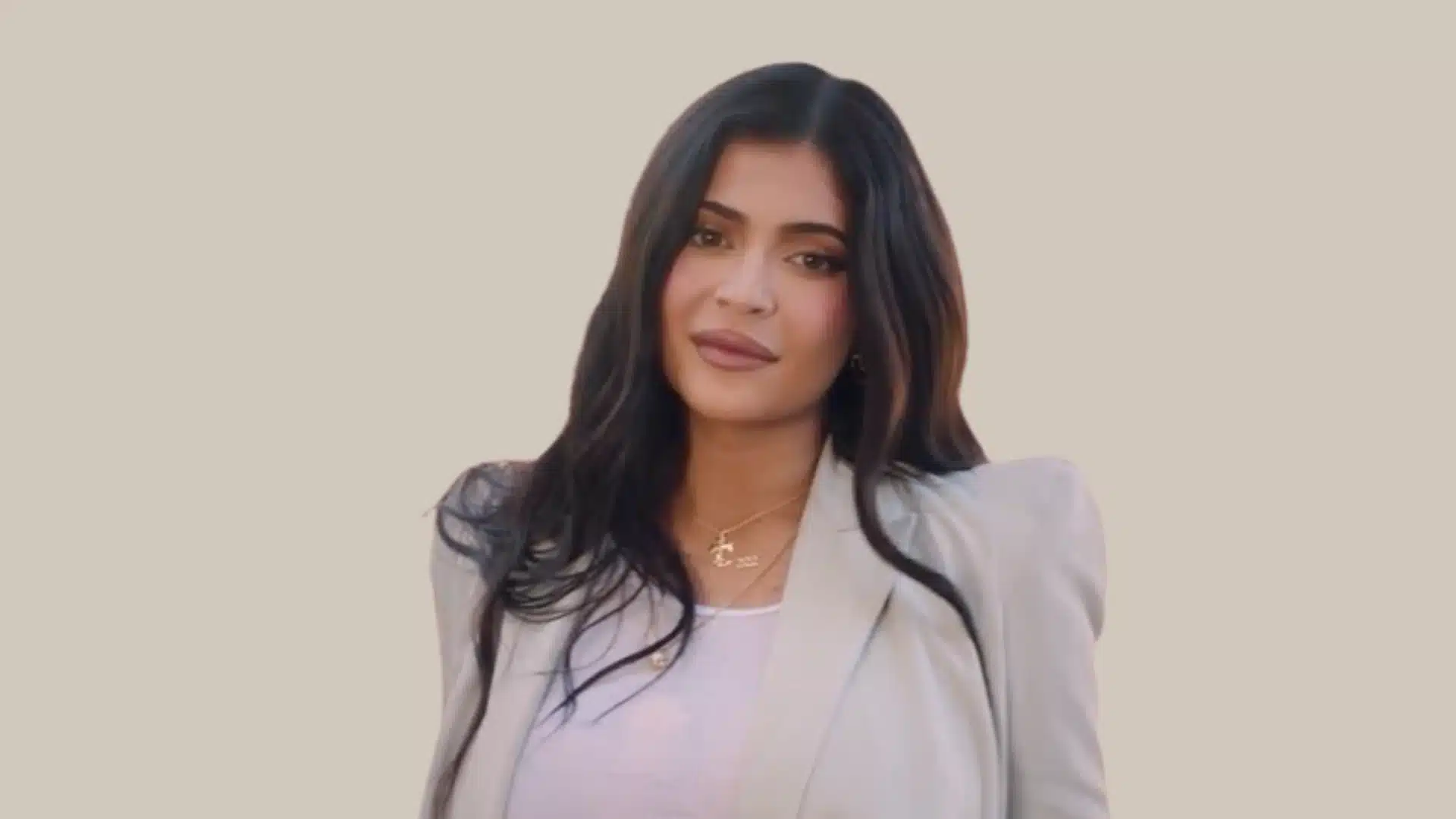 Kylie Jenner with long dark hair wearing a light blazer and white top against a solid beige background