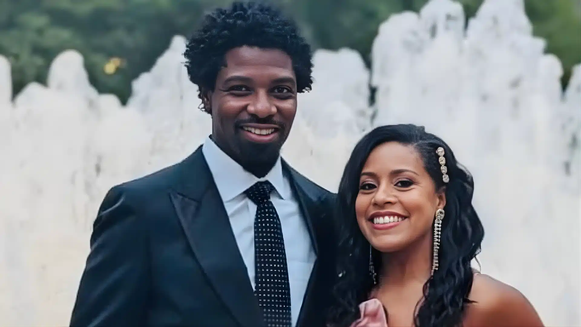 Uche Ojeh and Sheinelle Jones dressed formally pose while standing outdoors in front of a large, white water fountain