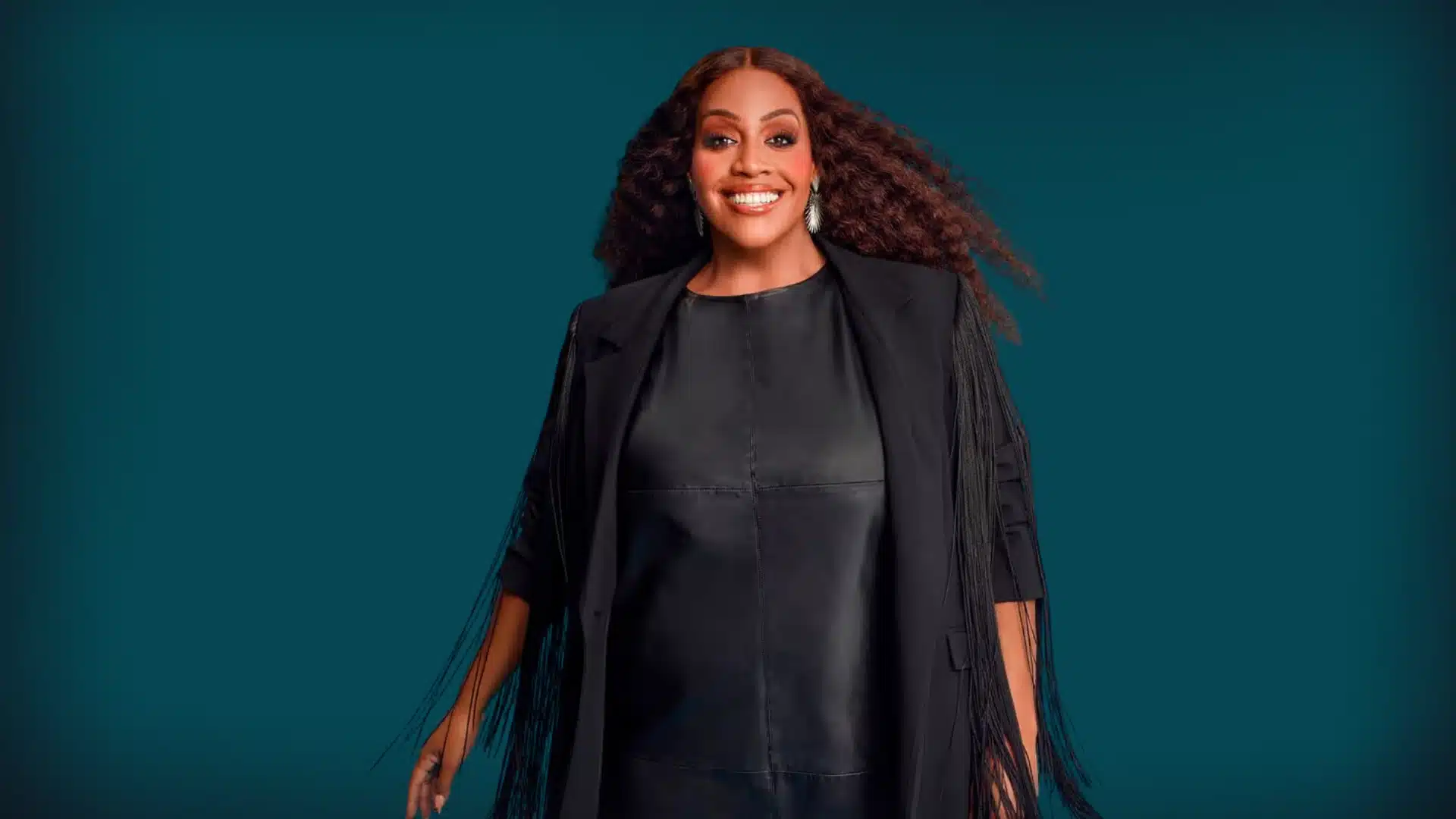 Alison Hammond, with voluminous auburn hair, wears a black leather dress and a fringed black jacket against a deep teal background