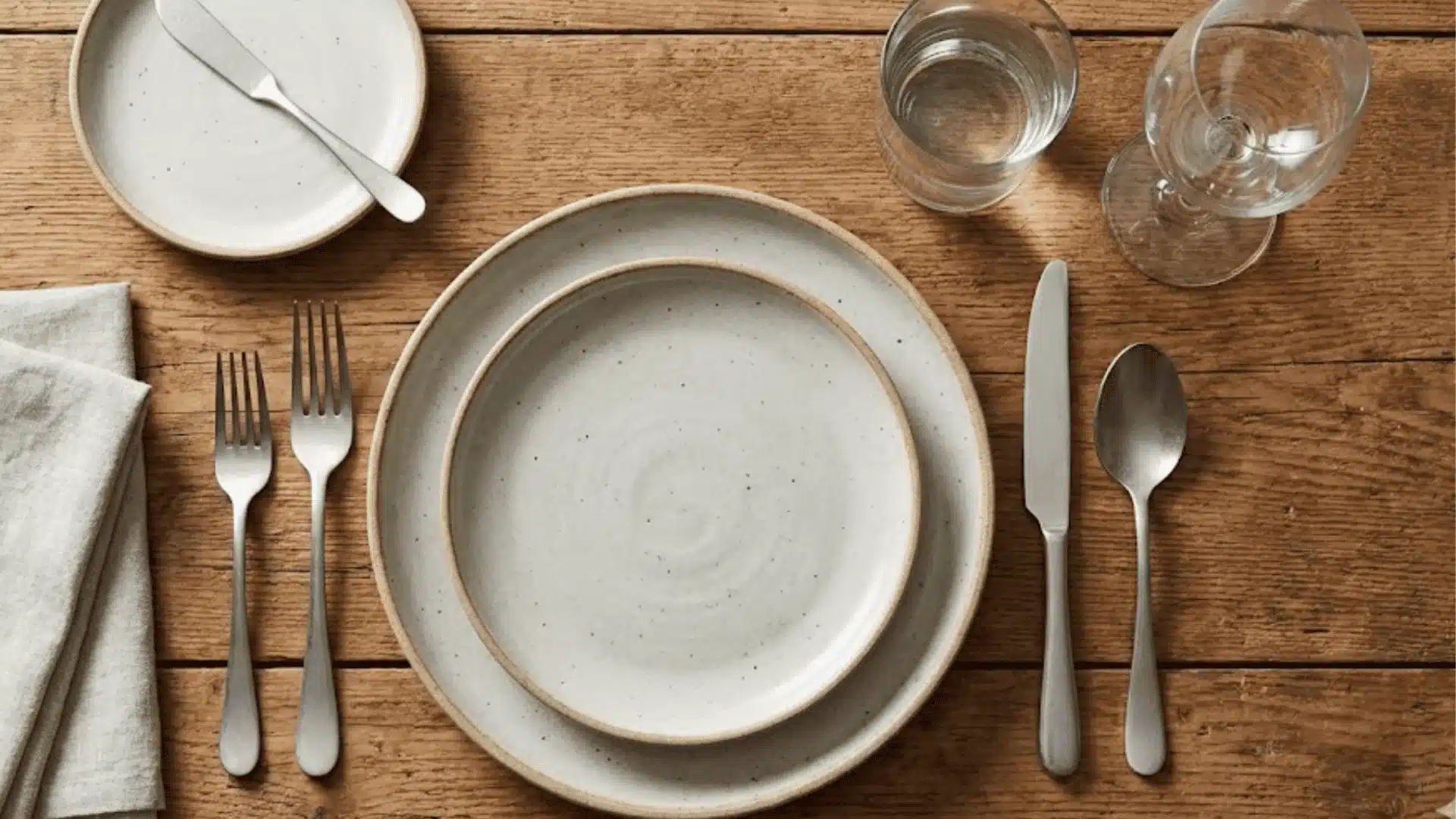 Casual table setting with speckled ceramic plates, silverware, glasses, and a linen napkin on a rustic wooden table