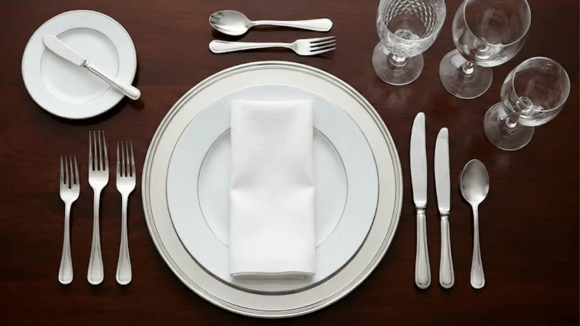 Formal table setting for one person, featuring stacked white plates, a folded white napkin, silverware arranged, and crystal glassware