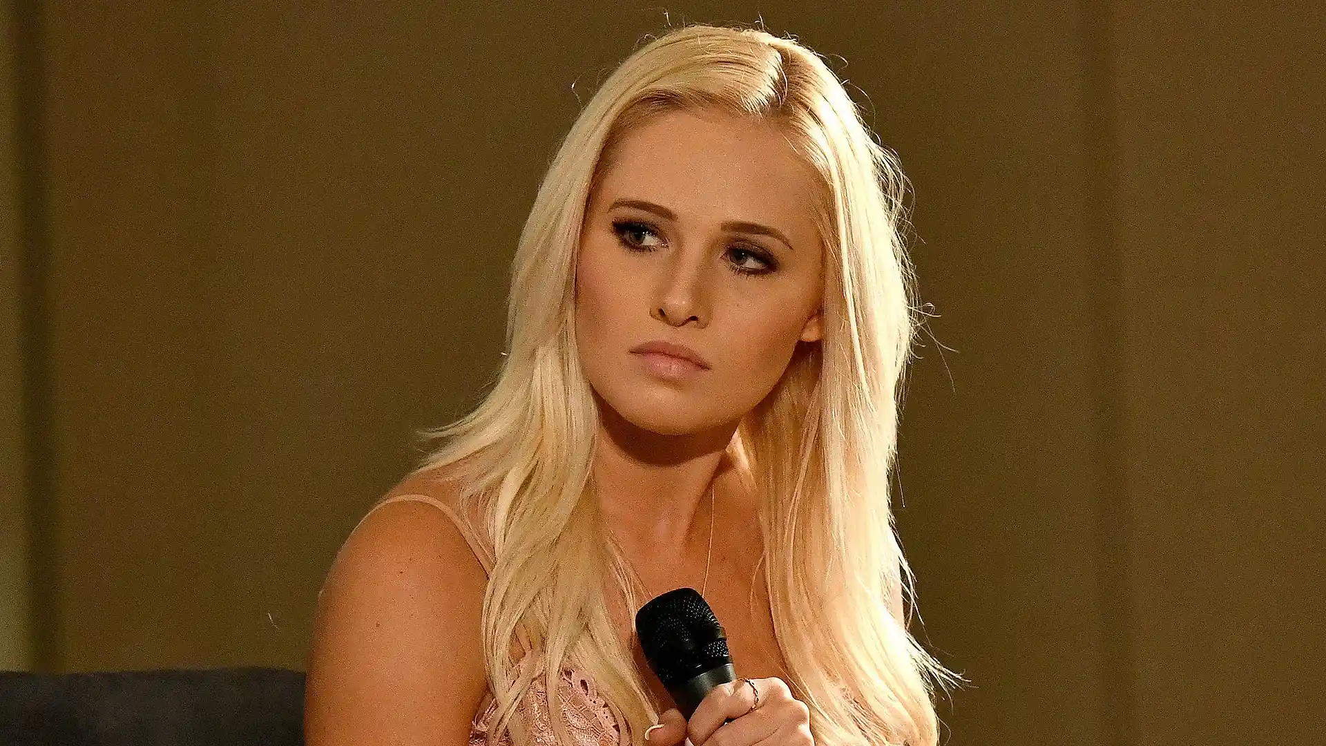 Tomi Lahren holds a black microphone against a solid olive-brown background