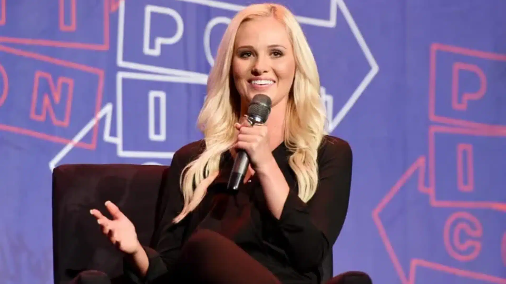 Tomi Lahren, in a black top, holding a microphone while seated on stage against a purple backdrop