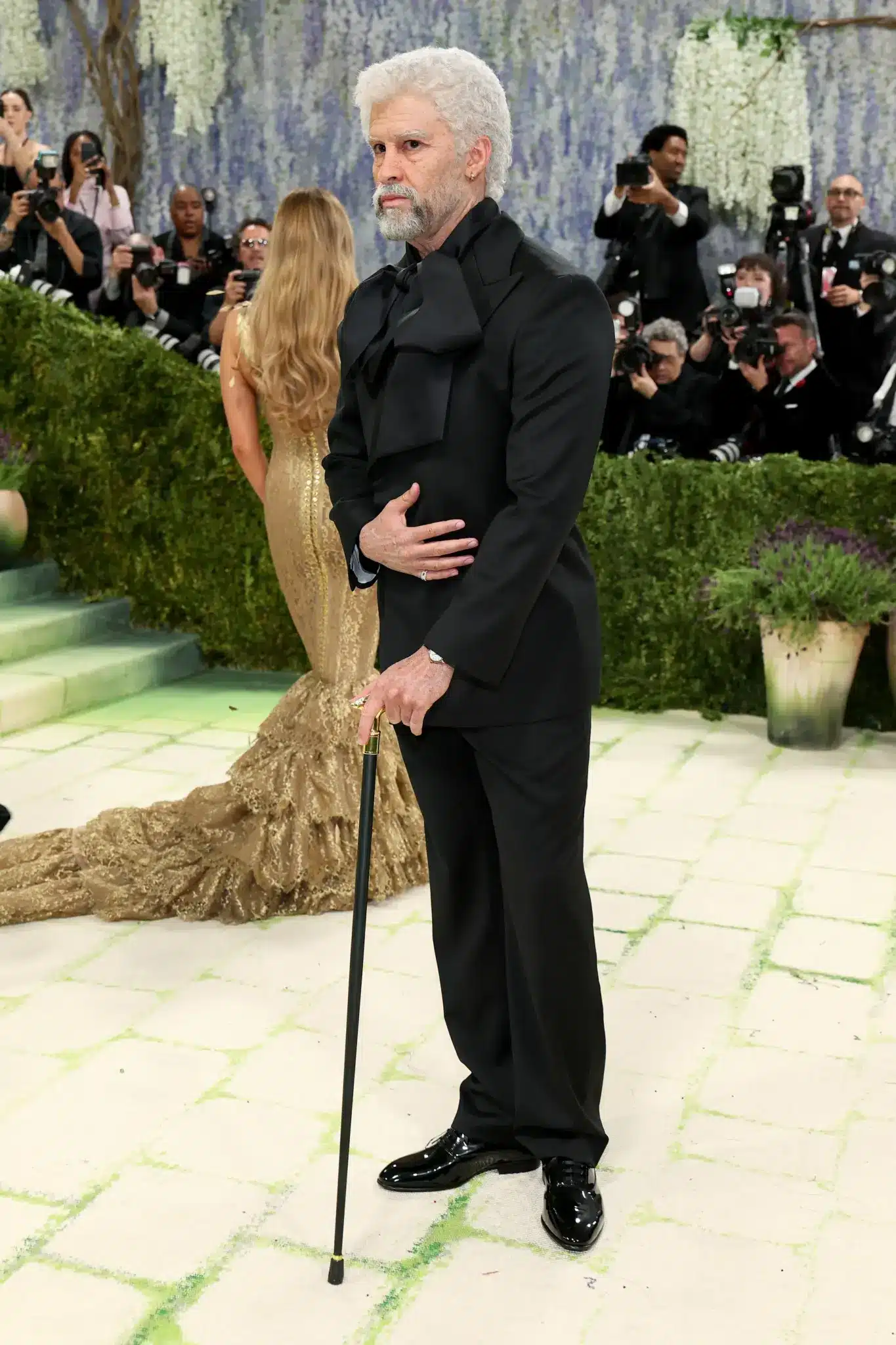 Bad Bunny poses in a black Zara suit using hyper-realistic prosthetics to age his face and hands while holding a cane