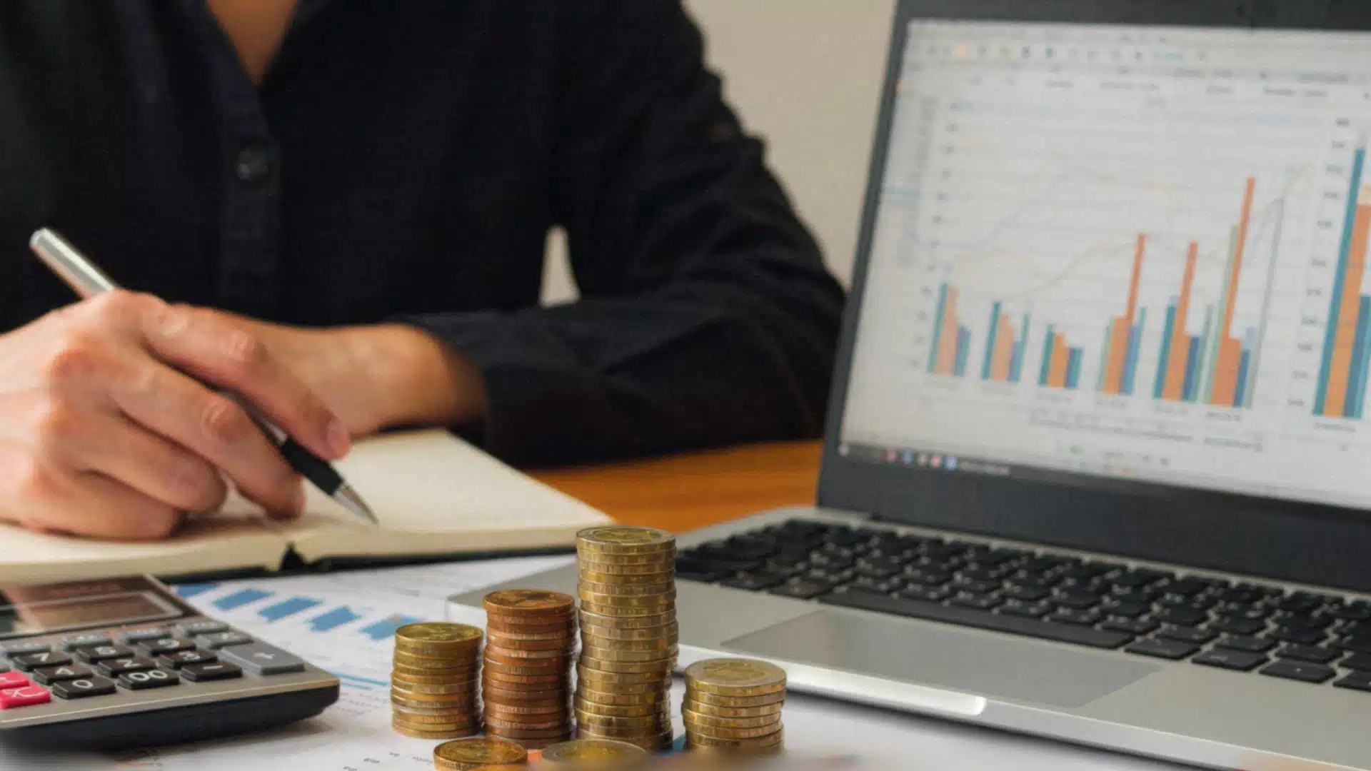 Person analyzing financial growth data on a laptop next to stacked coins, a calculator, and a notebook